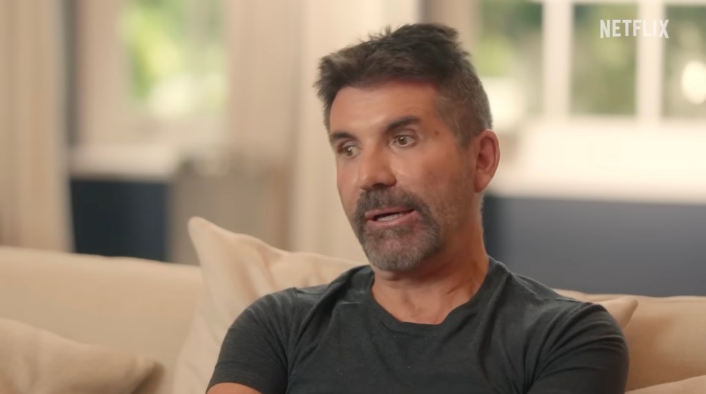 Simon Cowell wearing a gray t-shirt sitting on a couch.