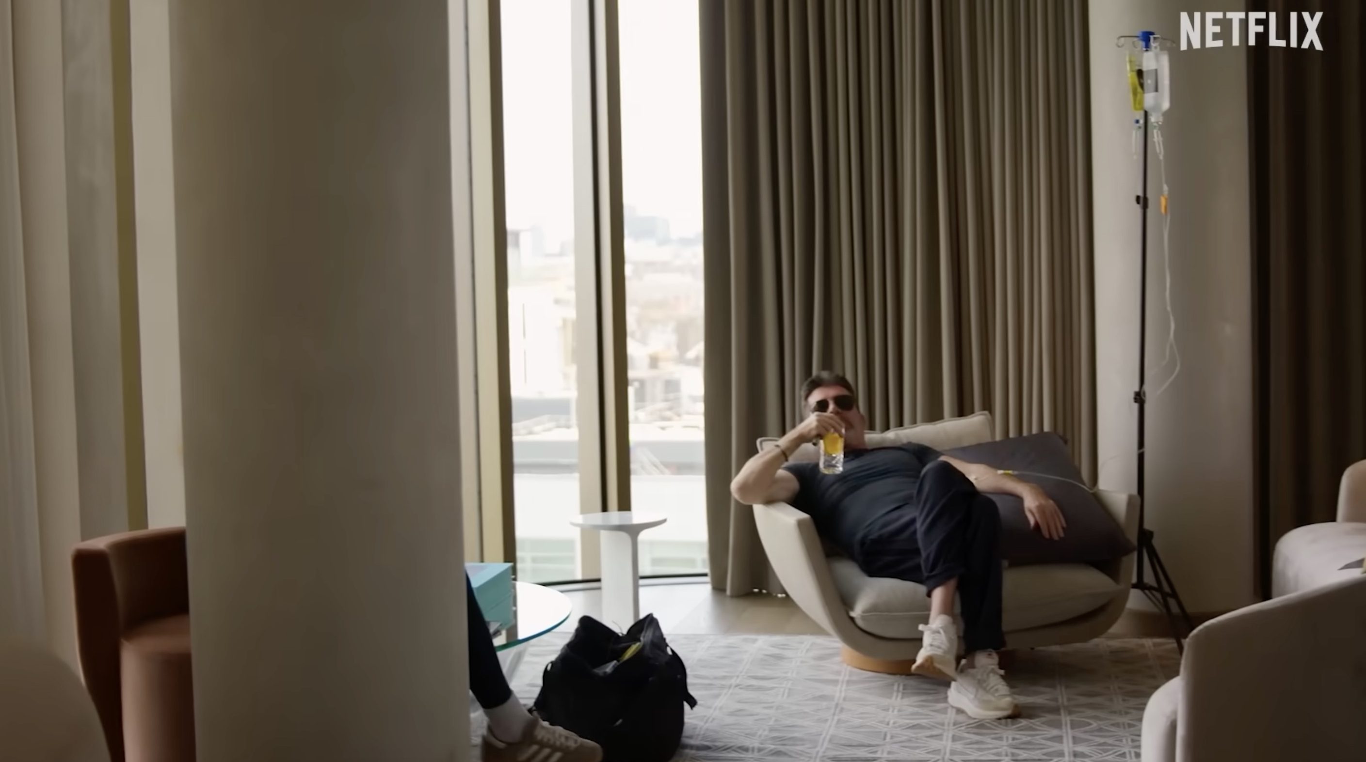 Simon Cowell, wearing sunglasses, a black T-shirt and black pants, sits in a chair drinking from a glass, with an IV drip pole next to him.