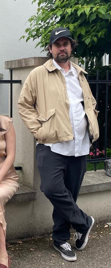 Nick Renier in a tan jacket, white shirt, black pants, black baseball cap, and black sneakers, leaning against a stone wall with a person in a satin dress seated next to him.