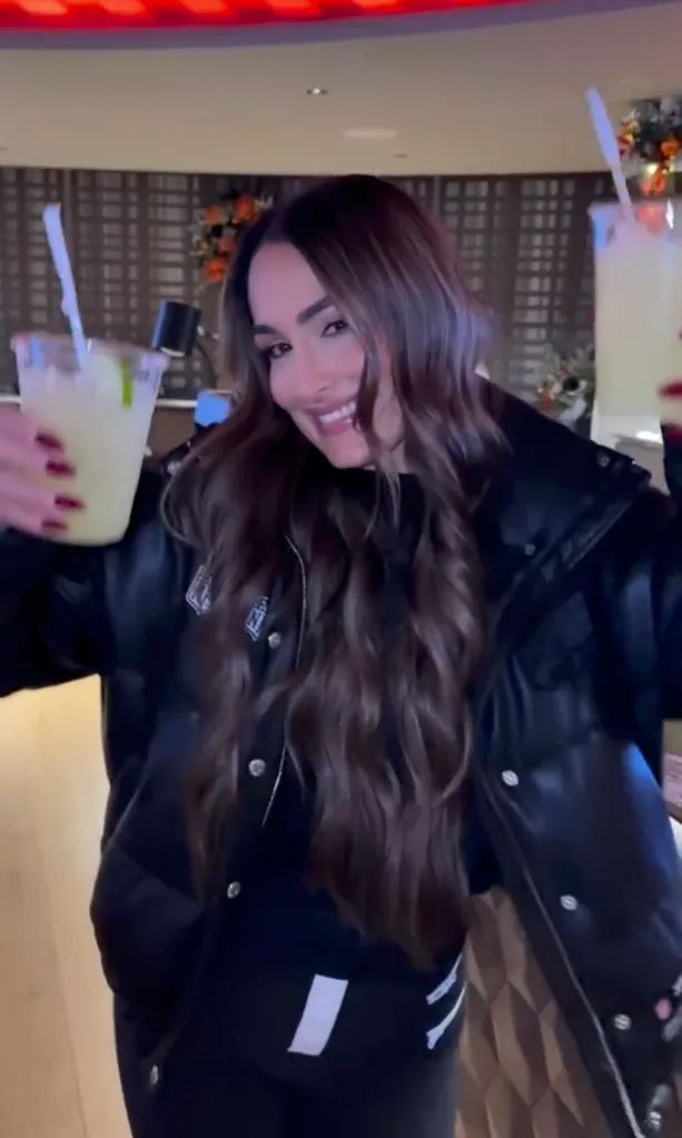 Nikki Bella in a black puffer jacket, holding two margaritas.