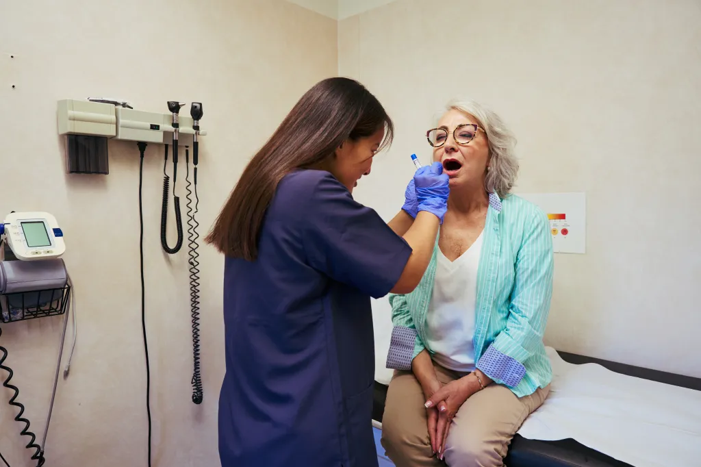 A nurse wearing gloves uses a tongue depressor to examine the throat of a senior woman during a medical checkup in a clinic.