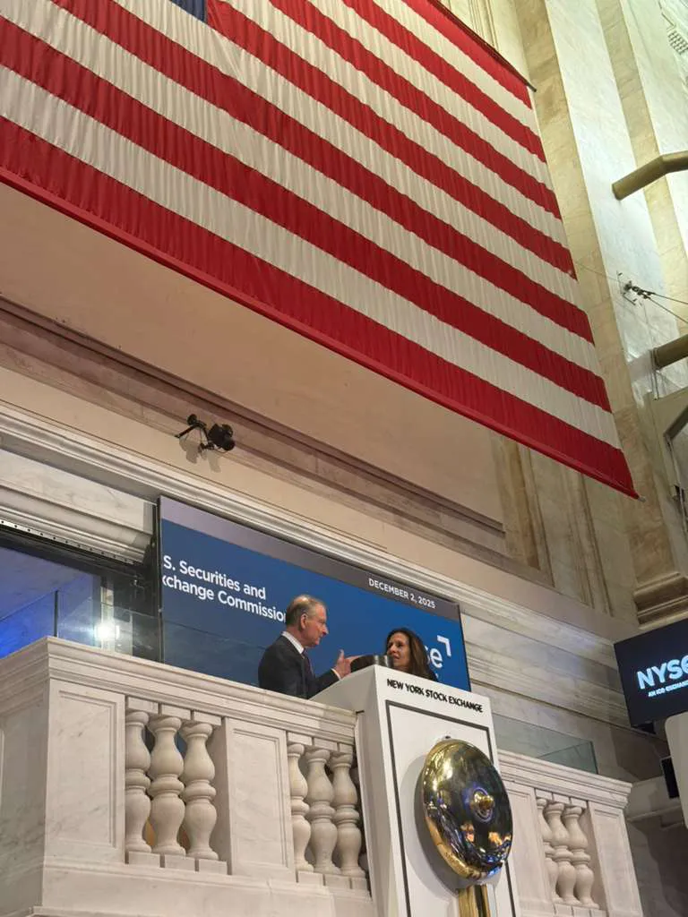 Two individuals speaking at a podium under a large American flag at the New York Stock Exchange.