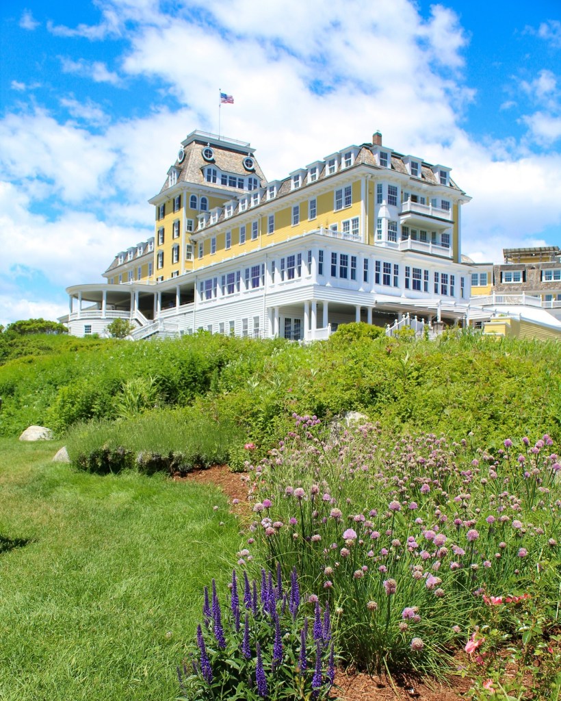 The Ocean House hotel in Rhode Island, a yellow building with white trim, viewed from a grassy hill with purple and pink flowers.