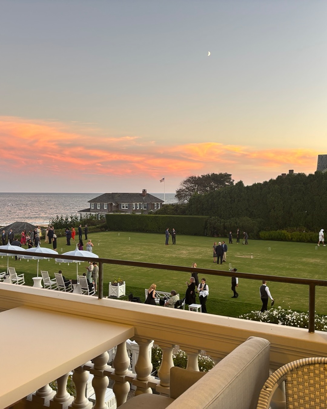 The Ocean House hotel and event space with people playing croquet on a lawn by the ocean at sunset.