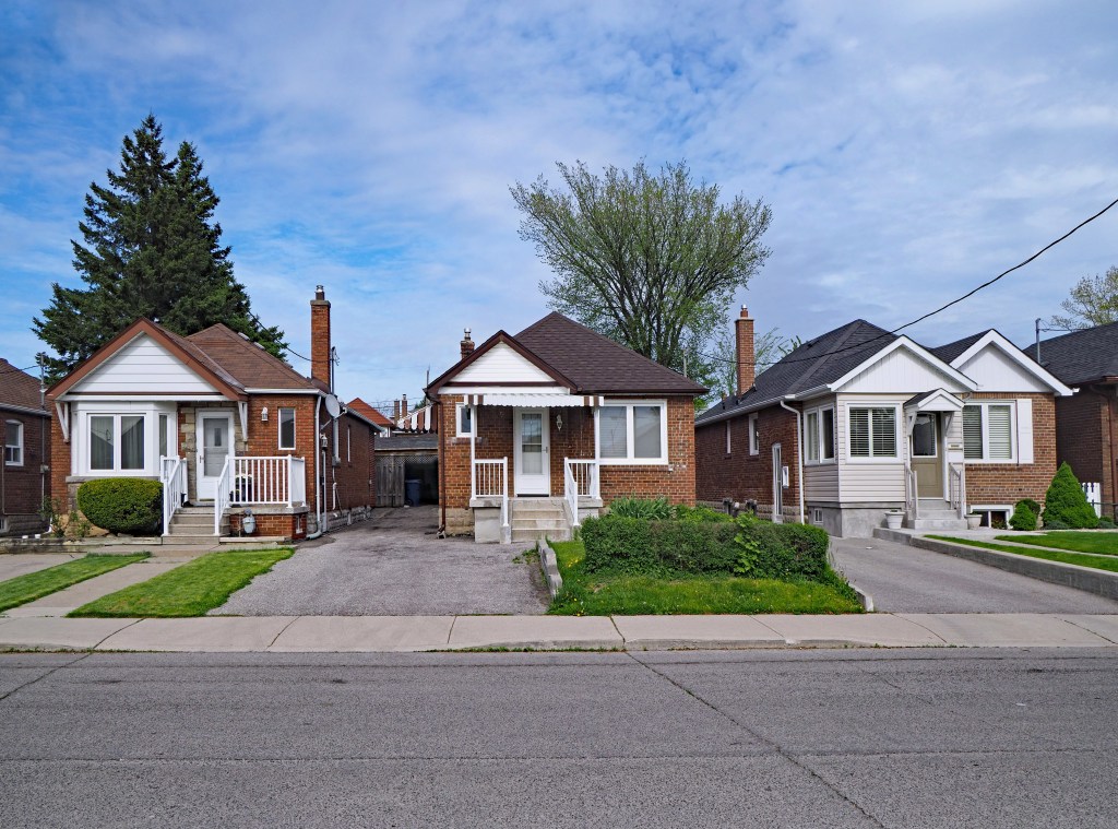 Row of three modest, 1950s-style brick bungalows with front yards and driveways under a cloudy sky.