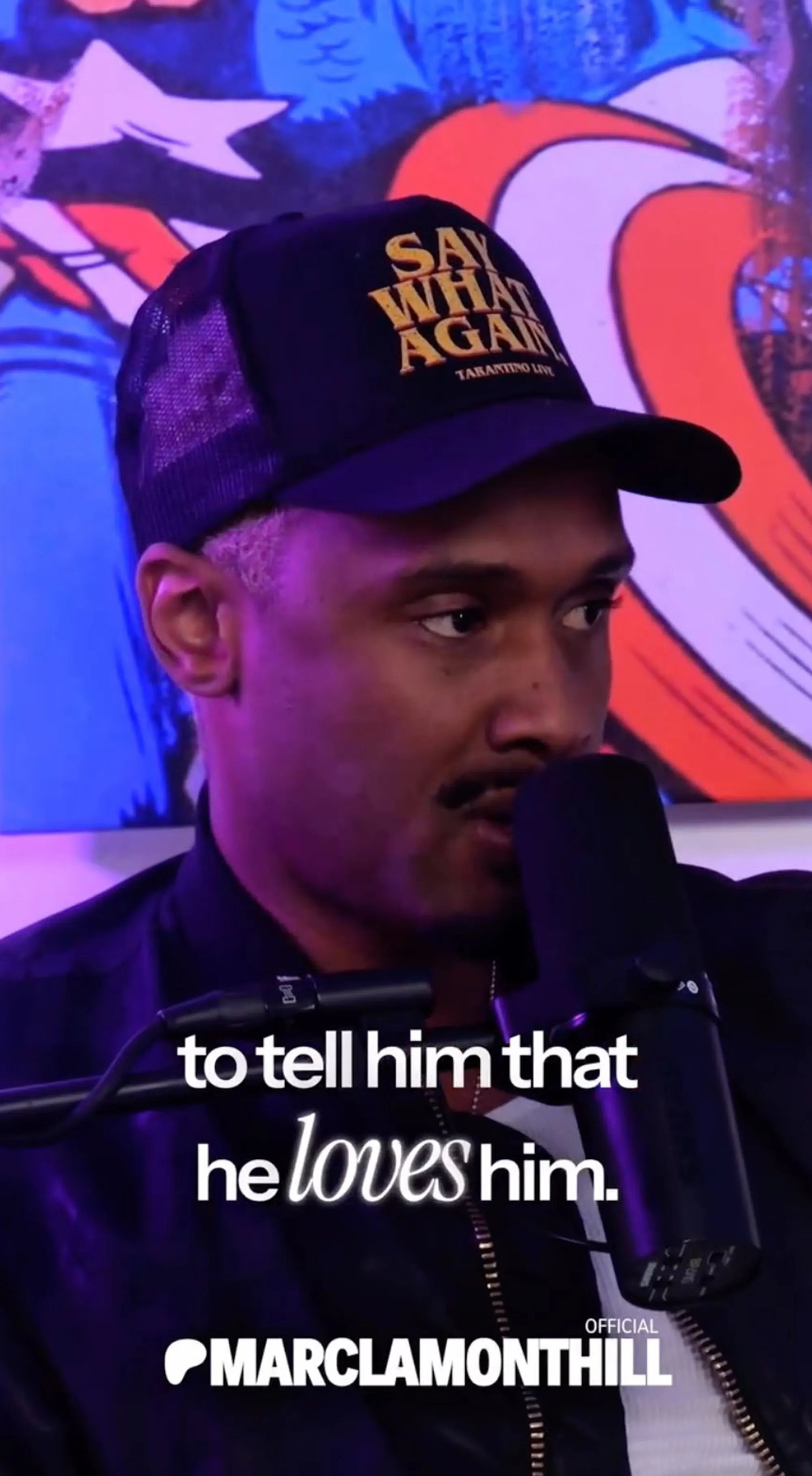 Brian McKnight Jr. in an interview with a microphone in front of him, wearing a black hat that says 