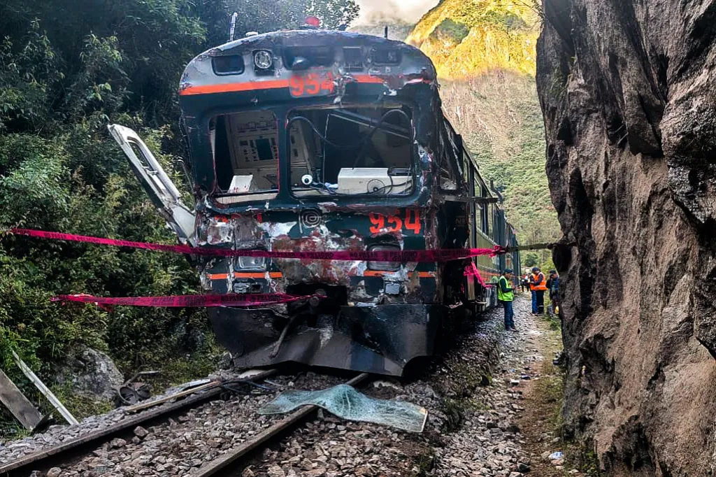 A severely damaged train, its front crumpled, rests on tracks with debris scattered around, as red caution tape marks off the area and a few people in safety vests stand nearby.