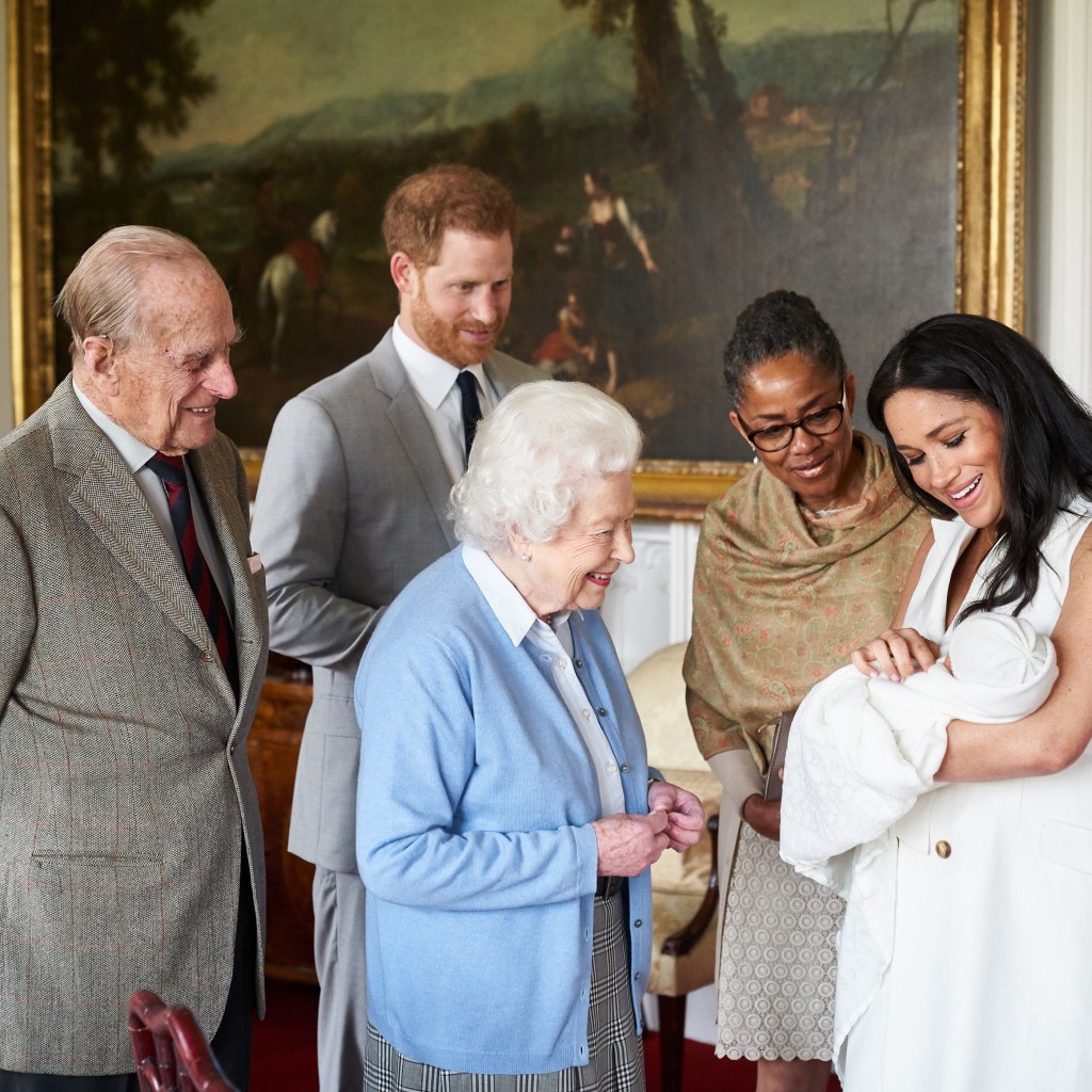 Prince Harry in a grey suit and white shirt and tie, and Duchess Meghan Markle, in a white dress, present their newborn son Prince Archie , in a white blanket, to Queen Elizabeth II , wearing a blue cardigan and white shirt and grey skirt, and Prince Philip, in a tweed jacket, white shirt and striped tie,, joined by Meghan's mother Doria Ragland, in a brown wrap.