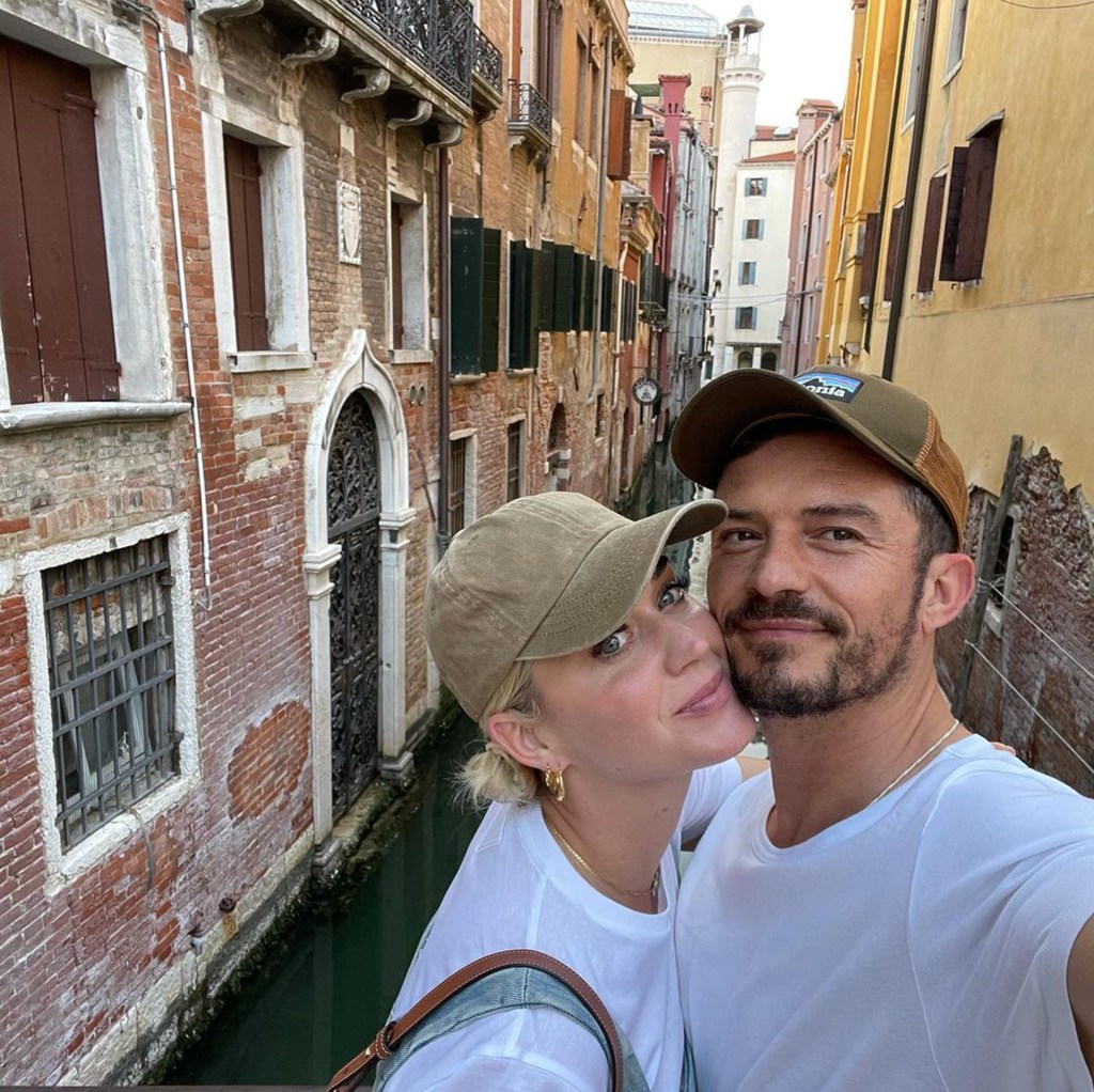 Katy Perry and Orlando Bloom smiling and wearing matching white t-shirts and caps while standing in front of a canal in Venice.