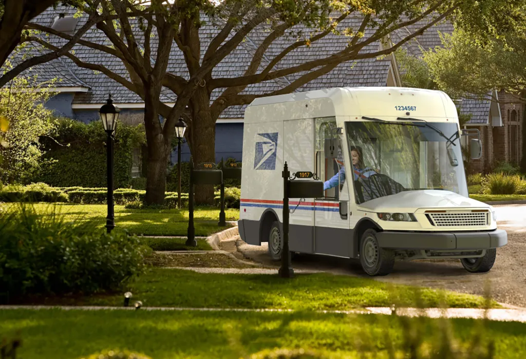 A mail carrier driving a white and gray Next Generation Delivery Vehicle on a residential street.