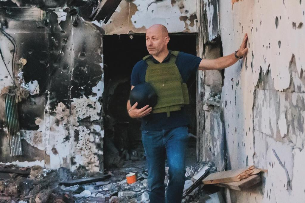 Arsen Ostrovsky, wearing a bulletproof vest and holding a helmet, stands in a damaged building at the site of the October 7 terror attack in Israel.