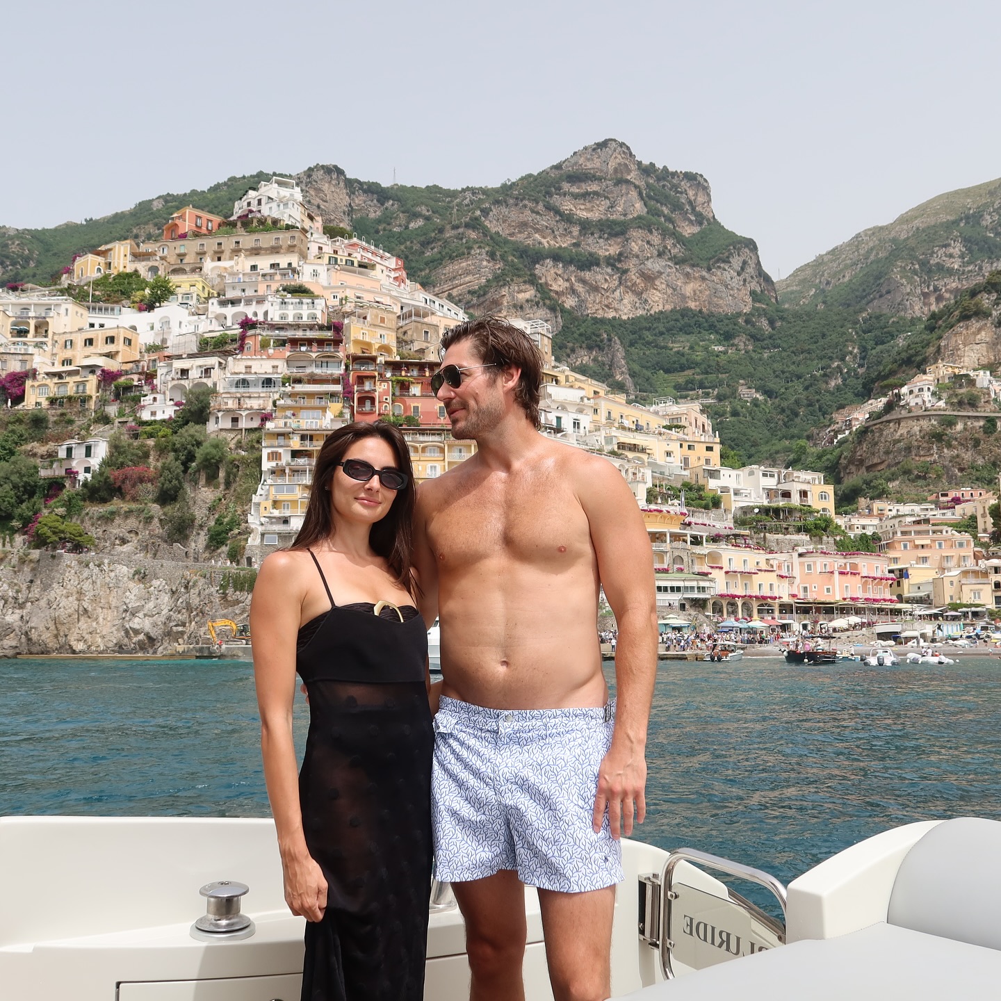 Paige DeSorbo and Craig Conover posing on a boat with the Positano coastline in the background.