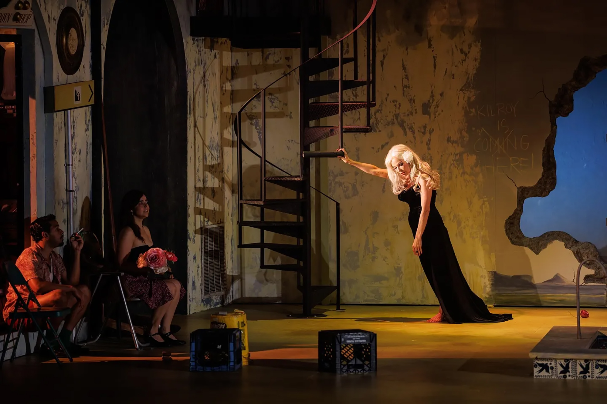 Pamela Anderson in a long black dress on a stage, holding onto a spiral staircase.