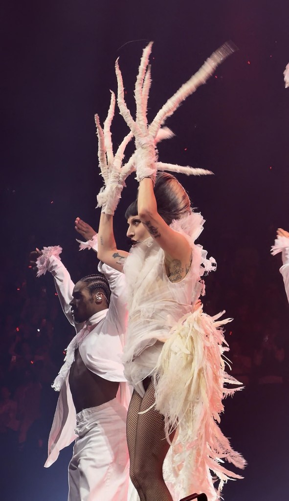 Lady Gaga performing in a white feathered and ruffled outfit and horned gloves at Accor Arena in Paris.