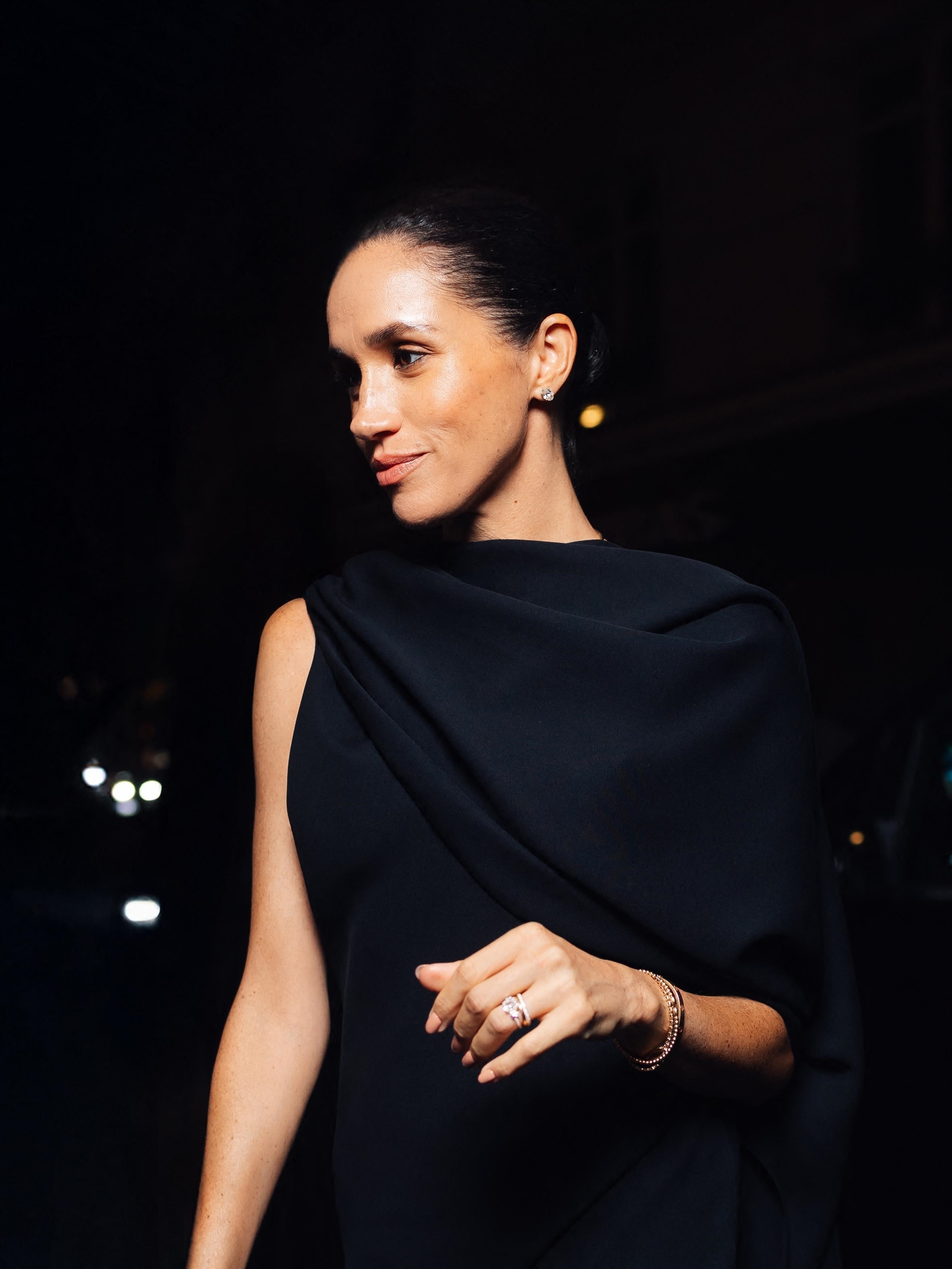 Meghan Markle, wearing a black dress with a draped neckline, a ring, and a bracelet, looking to the side.