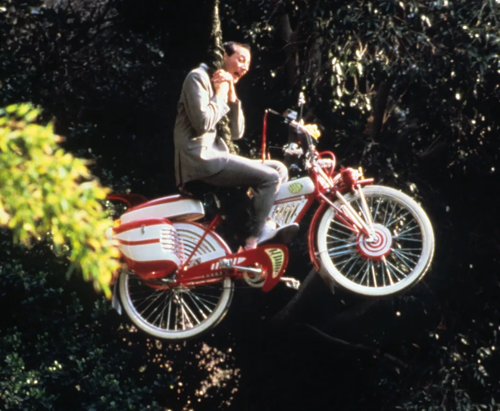 Pee-wee Herman holding onto a tree branch while flying on his red and white bicycle.