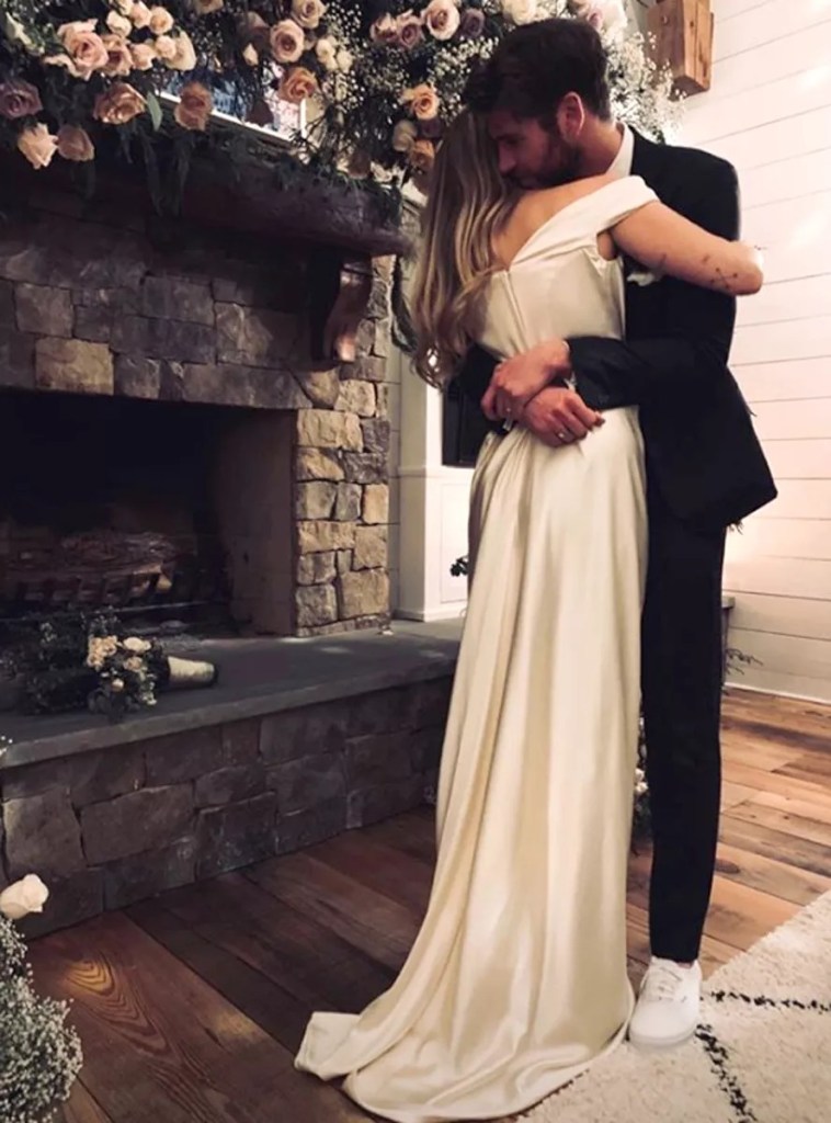 Miley Cyrus and Liam Hemsworth embracing at their wedding, standing in front of a stone fireplace decorated with flowers.