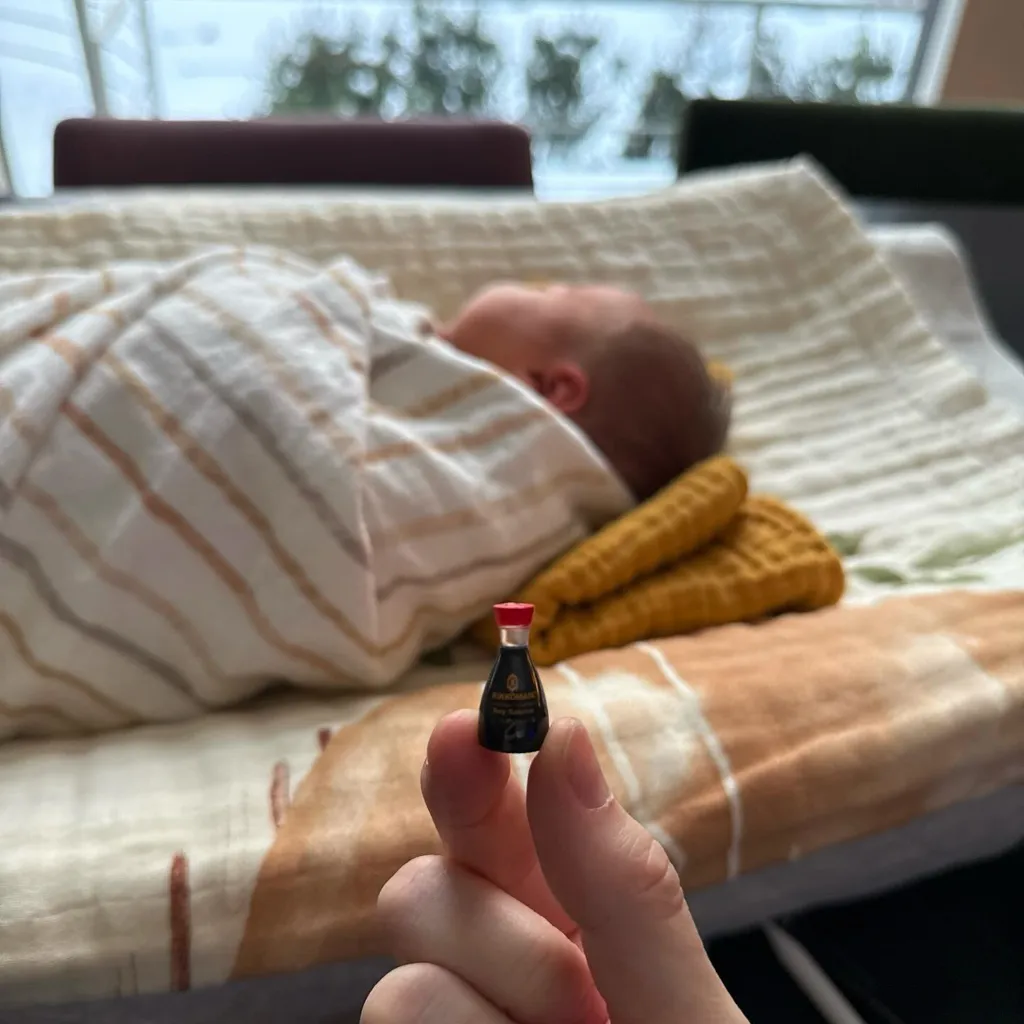 A hand holding a miniature Kikkoman soy sauce bottle in front of a baby swaddled in a blanket.