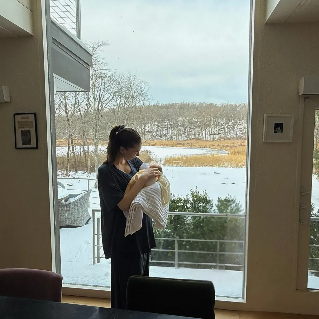 Elsie Hewitt holding her newborn baby while looking out a large window at a snowy landscape.