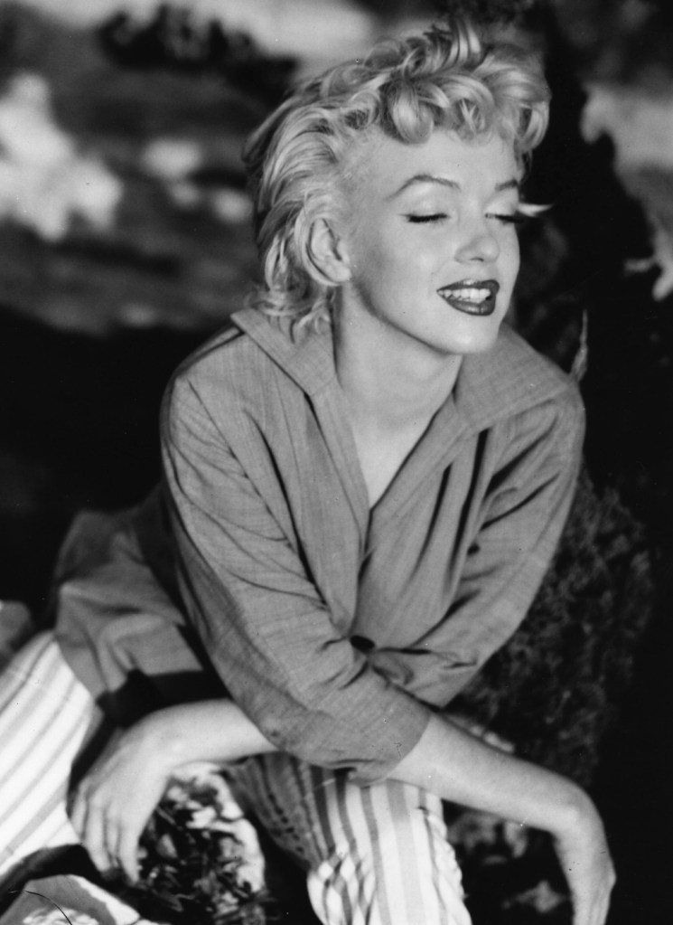 Marilyn Monroe, wearing a collared shirt and striped pants, smiles with her eyes closed.