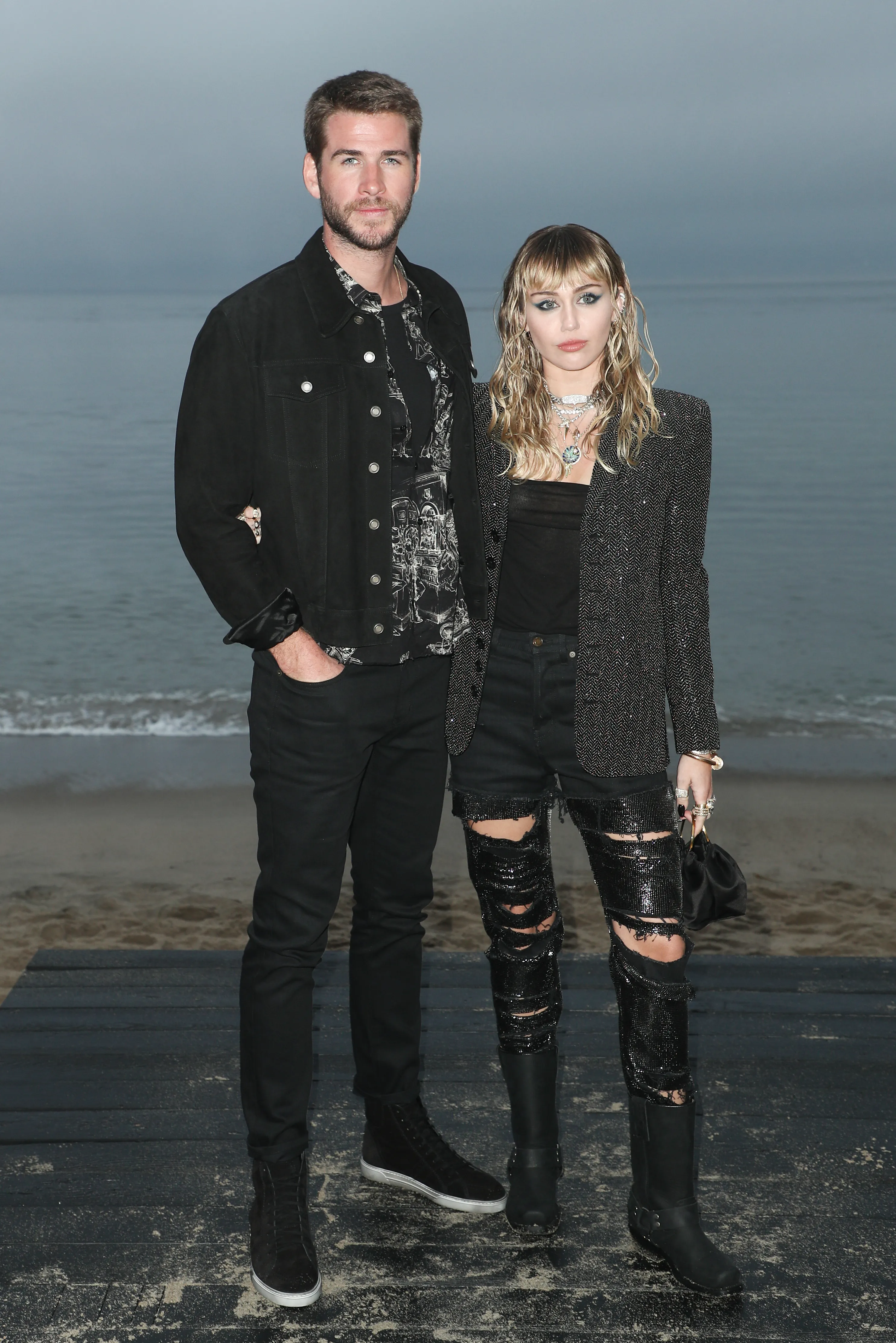 Liam Hemsworth and Miley Cyrus standing side-by-side on a wooden boardwalk at the beach.