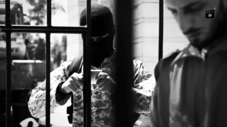 Black and white photo of a masked individual in camouflage and another man standing in a gated area, with an ISIS flag in the top right.