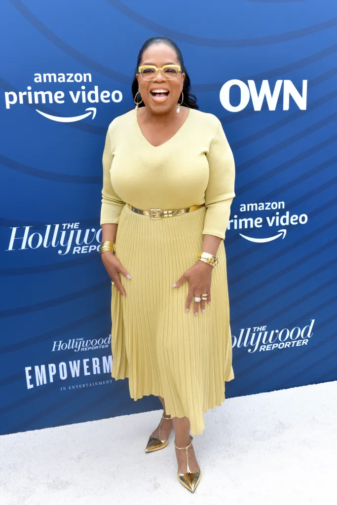 Oprah Winfrey in a yellow pleated dress with a gold belt, golden cuffs, and golden shoes, standing at The Hollywood Reporter's Empowerment In Entertainment Event 2019.