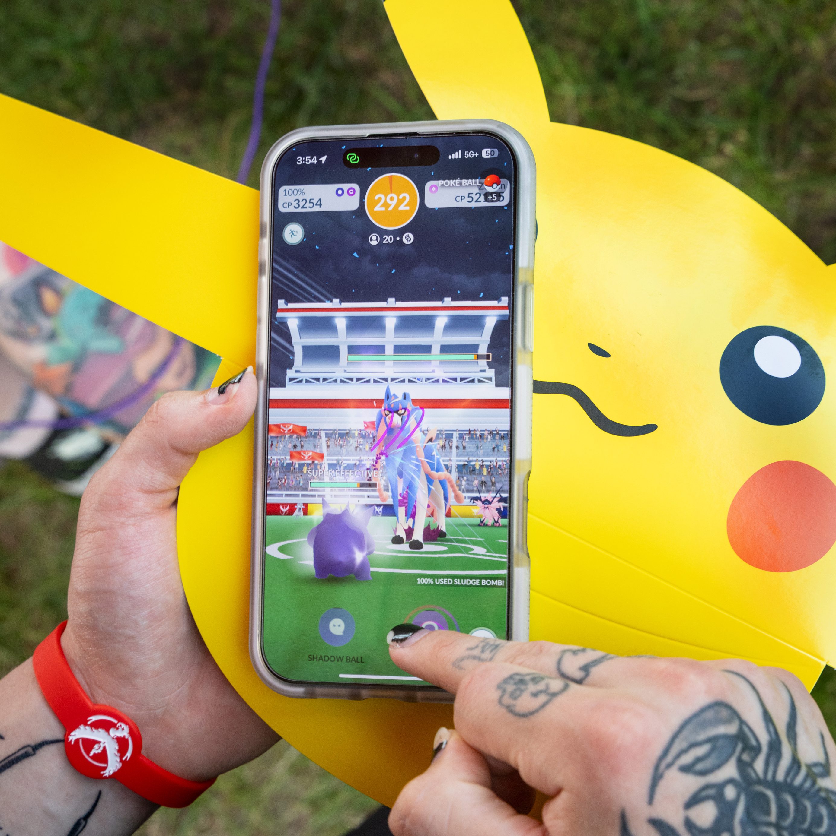 A trainer playing Pokémon GO on a smartphone, with a yellow Pikachu cutout behind the phone.