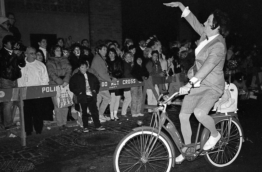 A person dressed as a female version of Pee-wee Herman on a bicycle, with a Pee-wee Herman doll on the back, waves to spectators during a parade.