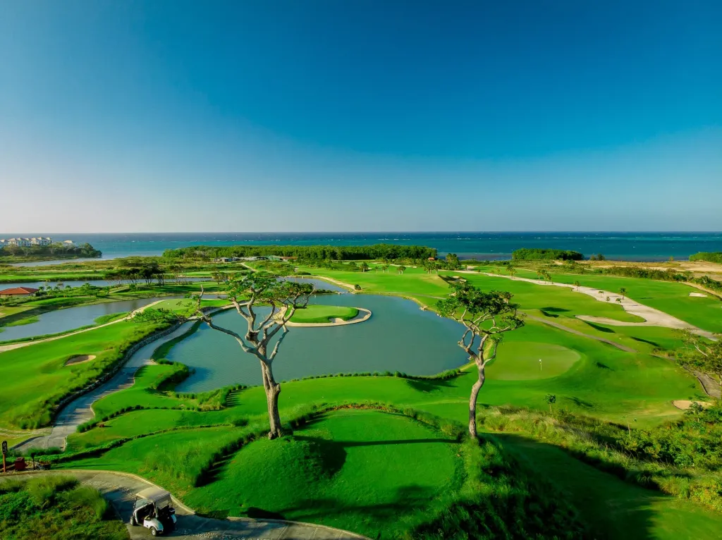 An aerial view of the 18-hole championship golf course in Roatán, with lush green fairways, water features, and the Caribbean Sea in the background.