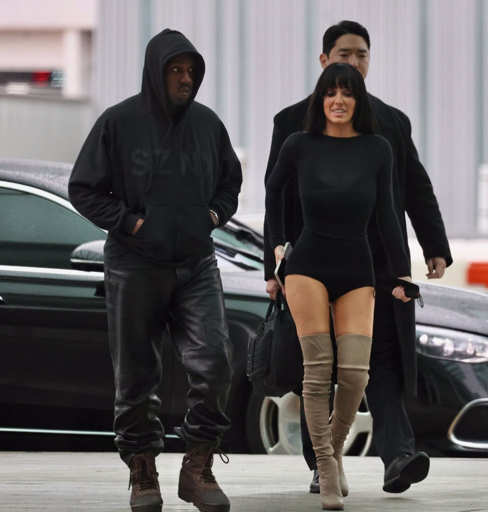 Kanye West in a black hoodie and leather pants, and Bianca Censori in a black bodysuit and beige over-the-knee boots, walk at Seoul Airport.