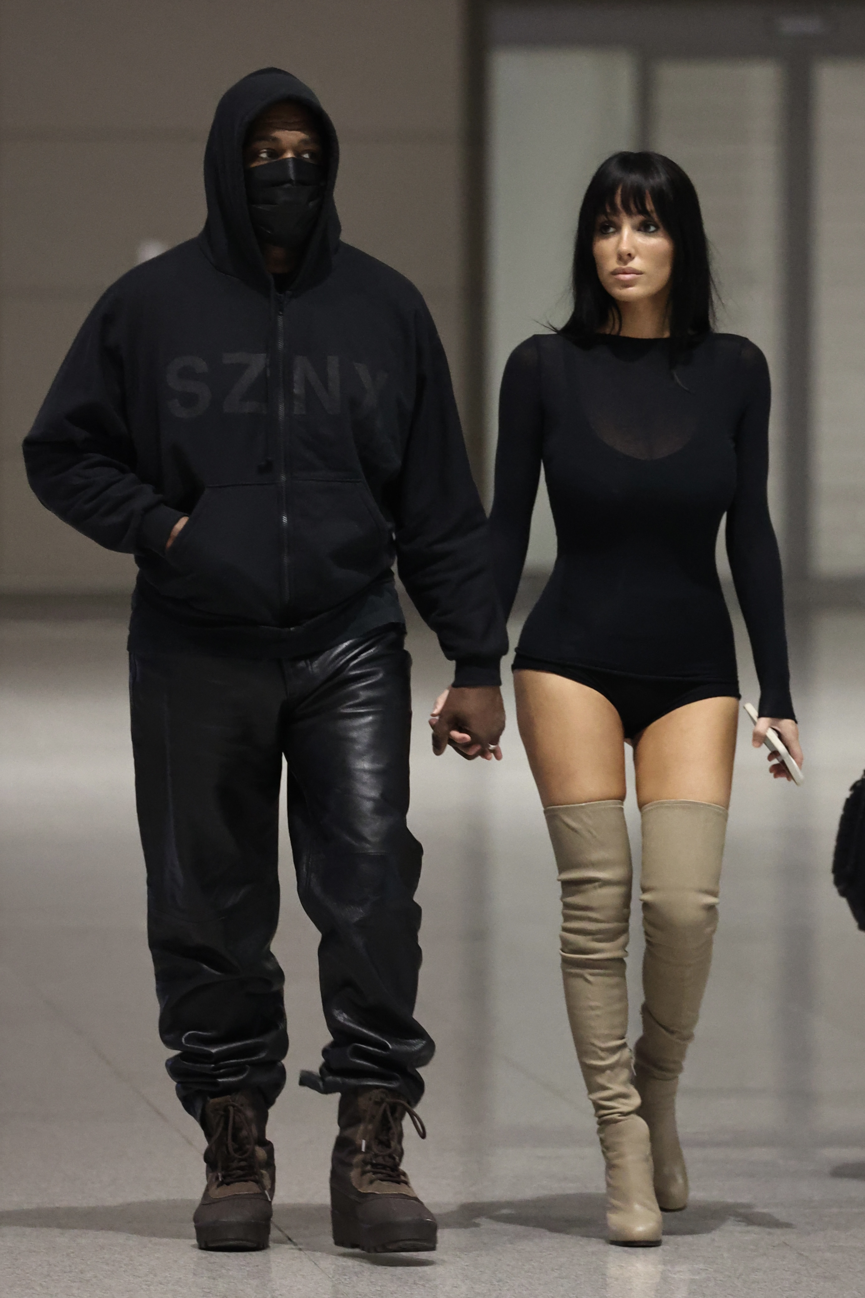 Kanye West in a black hoodie and leather pants, holding hands with Bianca Censori in a black bodysuit and beige thigh-high boots, at Seoul Airport.