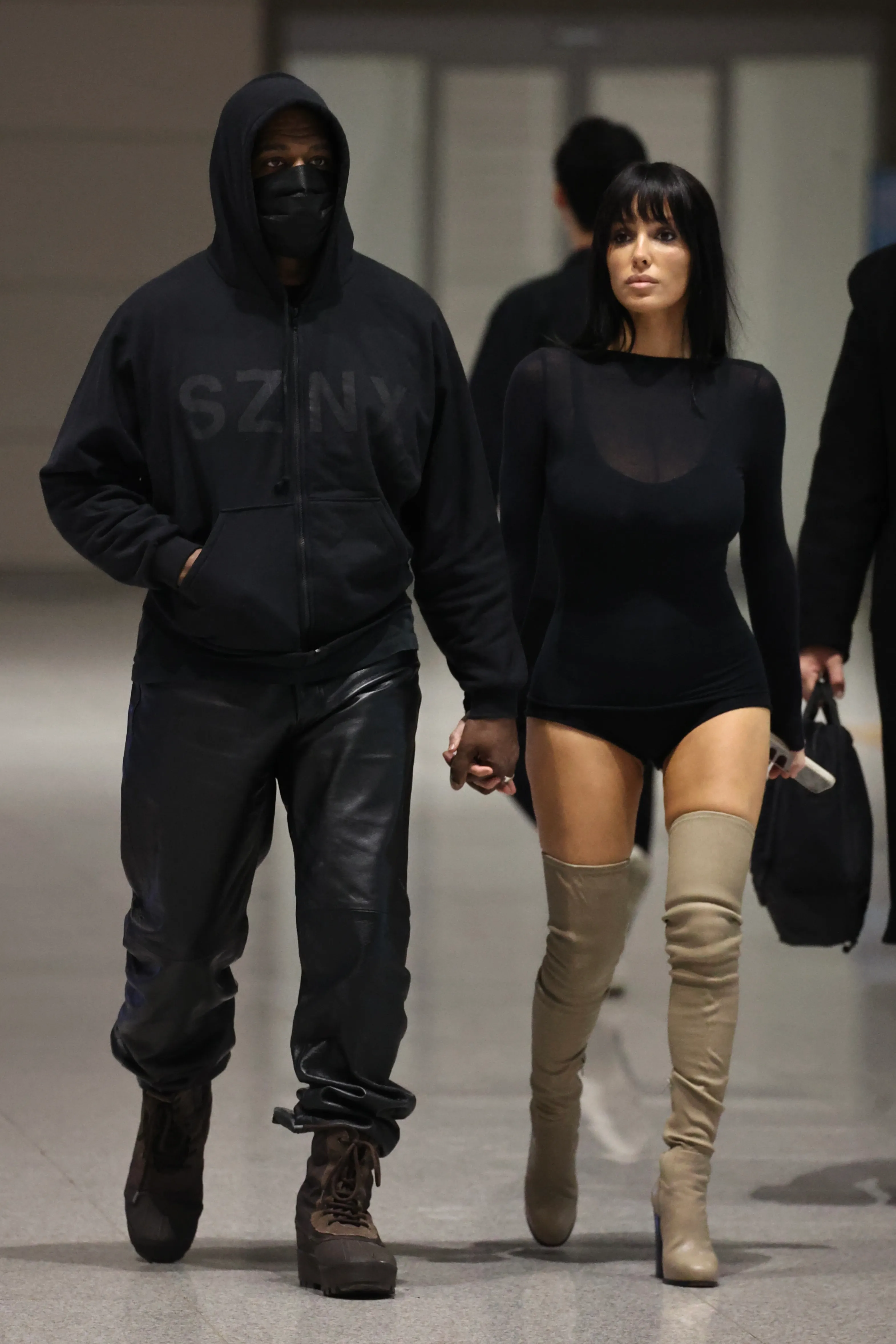 Kanye West in a black hoodie and mask holding hands with Bianca Censori in a black bodysuit and beige thigh-high boots at Seoul Airport.