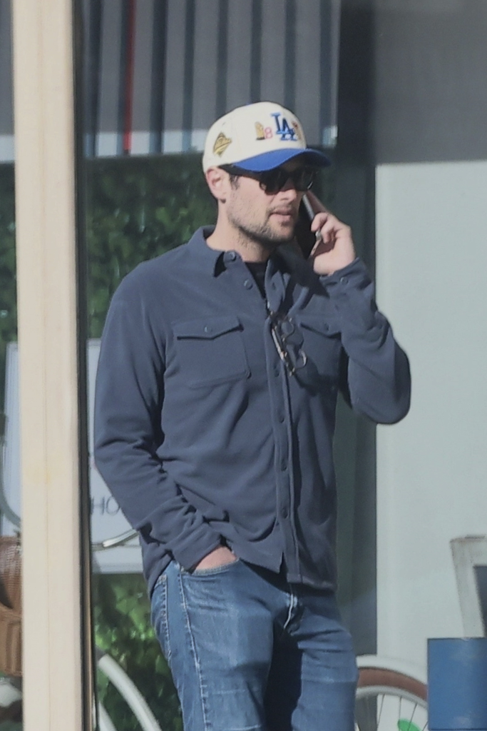 Jake Reiner wearing a Los Angeles Dodgers hat, blue long-sleeve shirt, and blue jeans while on the phone.