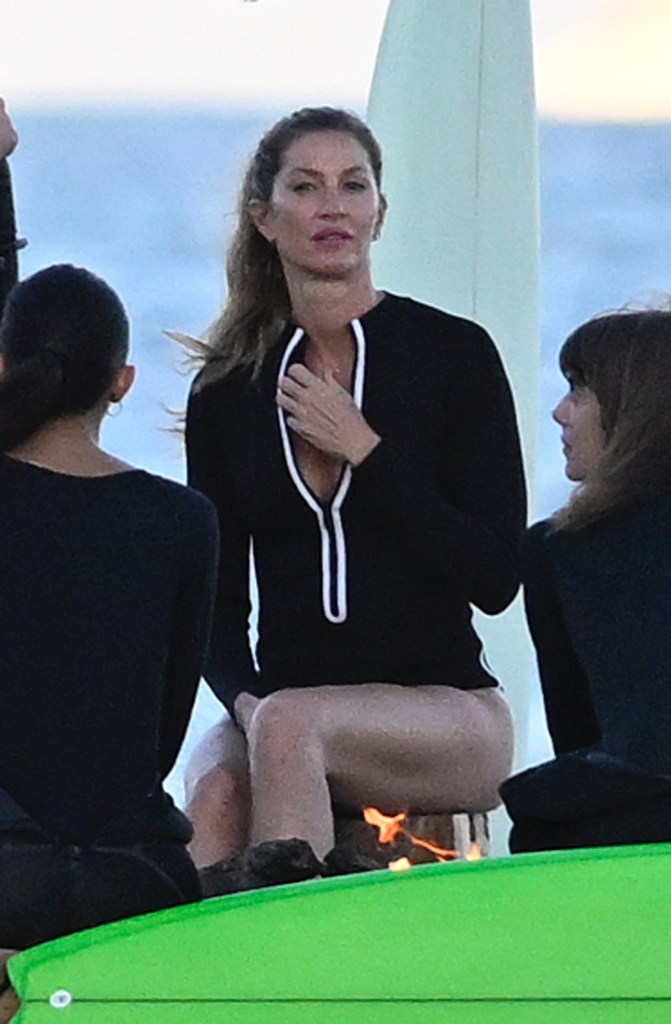 Gisele Bündchen sitting with a surfboard and a small fire.