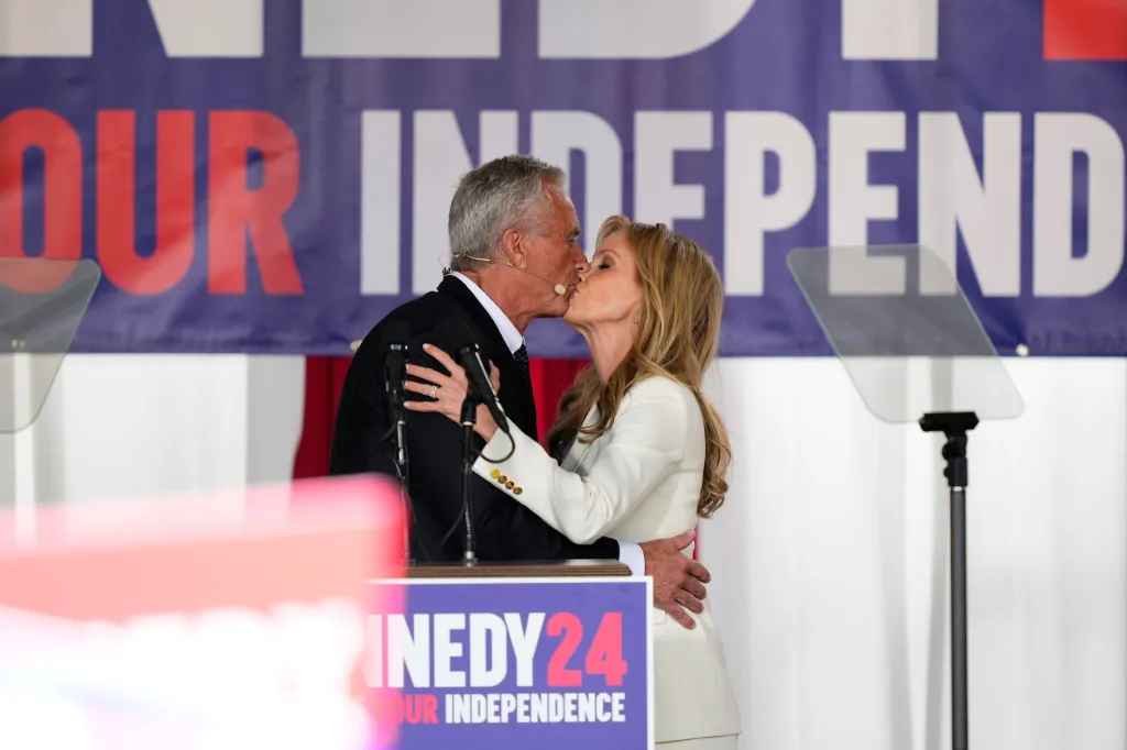 Robert F. Kennedy, Jr. in a black suit, and his wife, Cheryl Hines, in a white suit, kiss at a campaign event.
