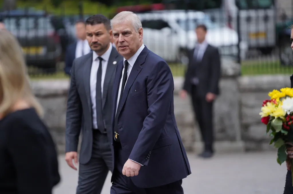 Prince Andrew in a navy suit and white shirt with a dark tie, looking towards the right, with a security guard behind him.