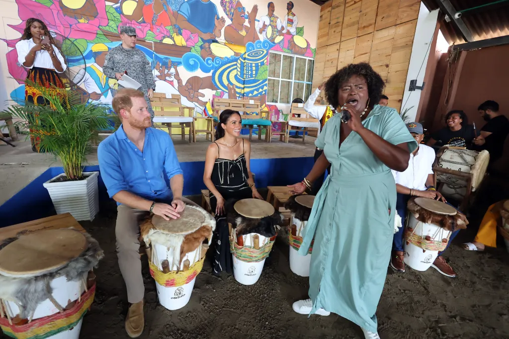 Prince Harry and Meghan, Duchess of Sussex, sitting at drums and watching a woman sing into a microphone.