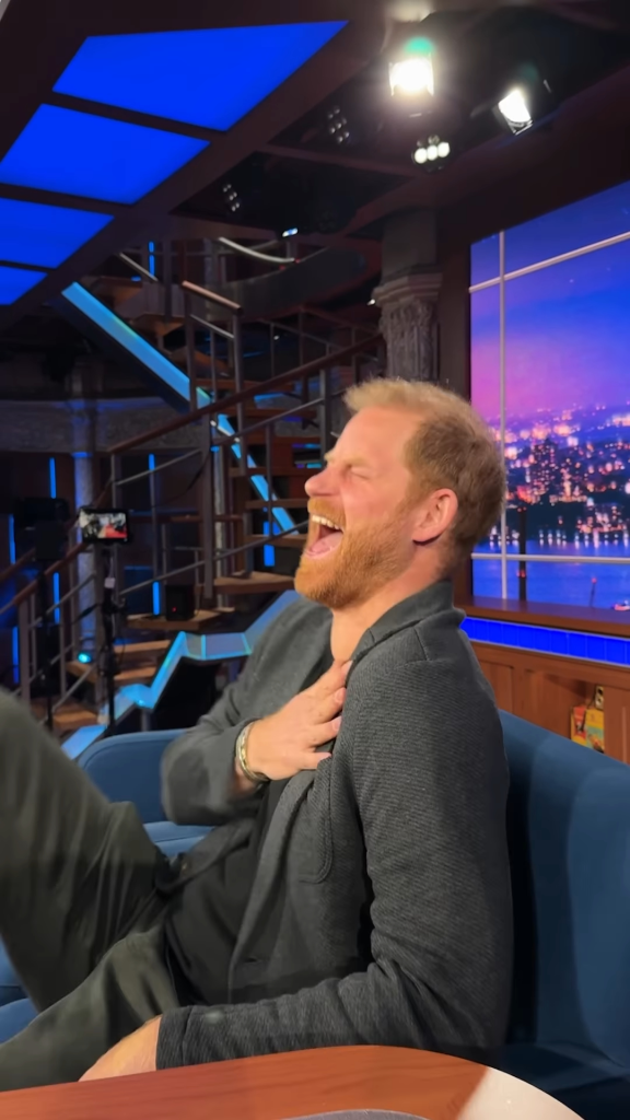 Prince Harry laughing boisterously on a blue couch during an interview.