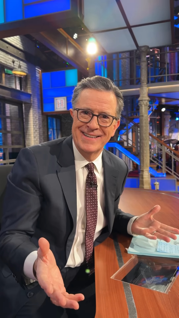 Stephen Colbert seated at a desk, smiling with open hands.