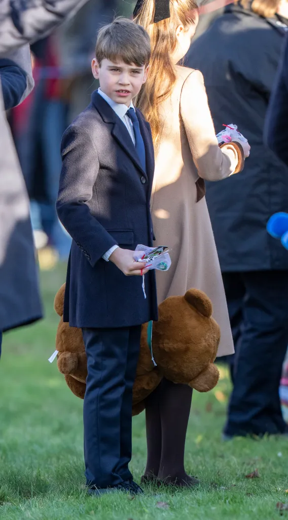 Prince Louis of Wales wearing a coat and tie, holding a small white bag, and carrying a large teddy bear.
