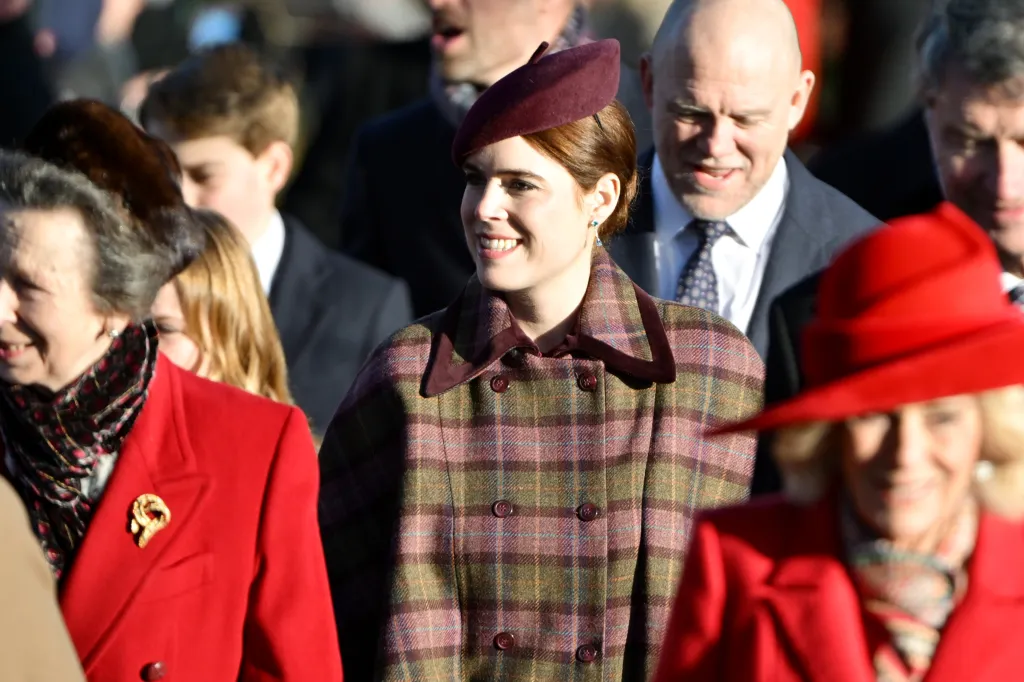 Princess Eugenie of York, wearing a plaid coat and a maroon beret, smiles as she walks among other members of the British Royal Family.