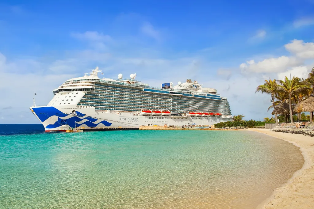 The Royal Princess cruise ship docked at a tropical beach with clear blue water and palm trees.