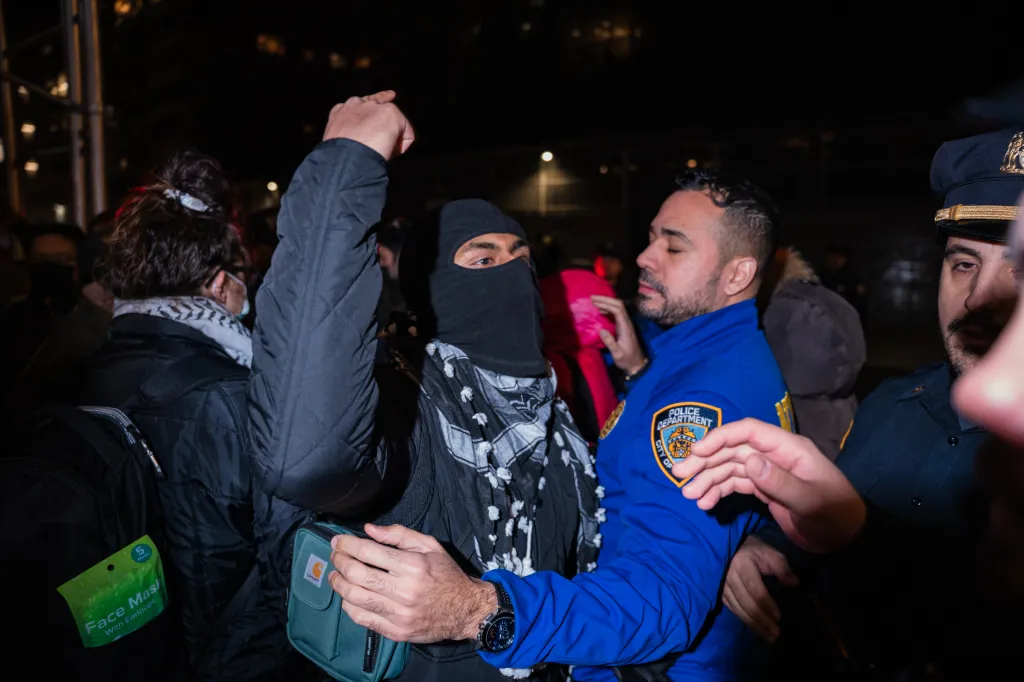 A pro-Palestinian protester with a balaclava and keffiyeh raises their fist while a police officer stands nearby during a demonstration outside a synagogue.