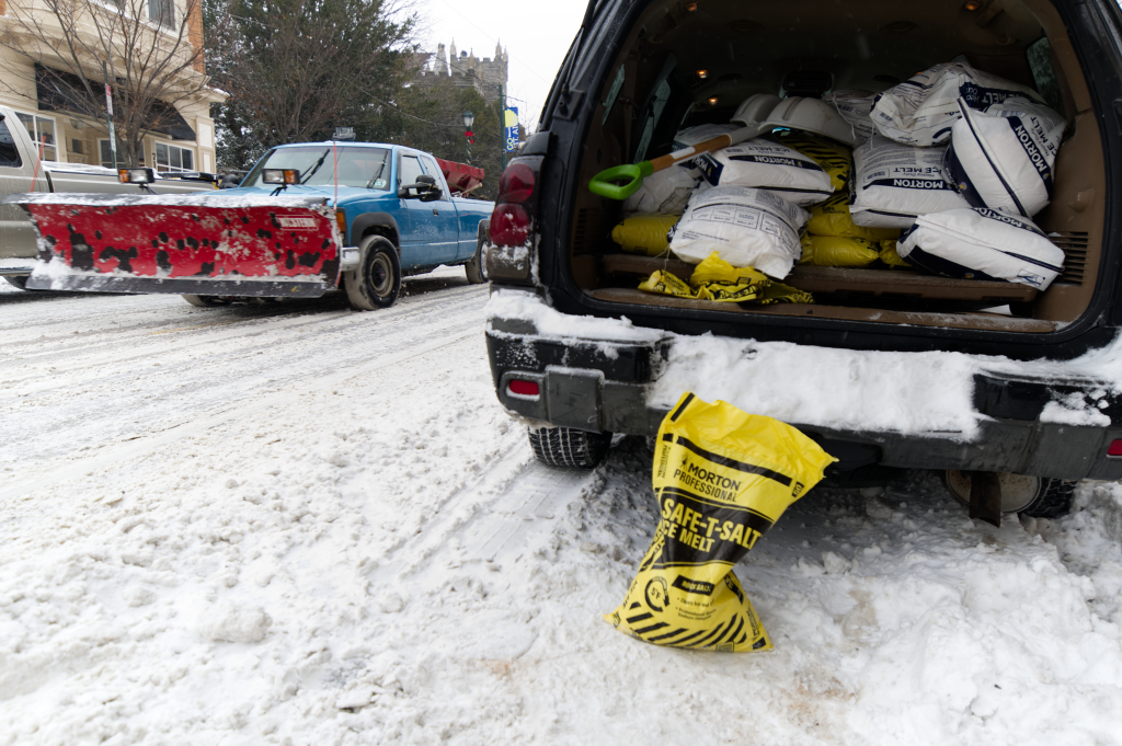 A blue pickup truck with a red snowplow and an SUV with its trunk open, full of bags of Morton Professional Safe-T-Salt ice melt, are parked on a snow-covered street in Philadelphia.