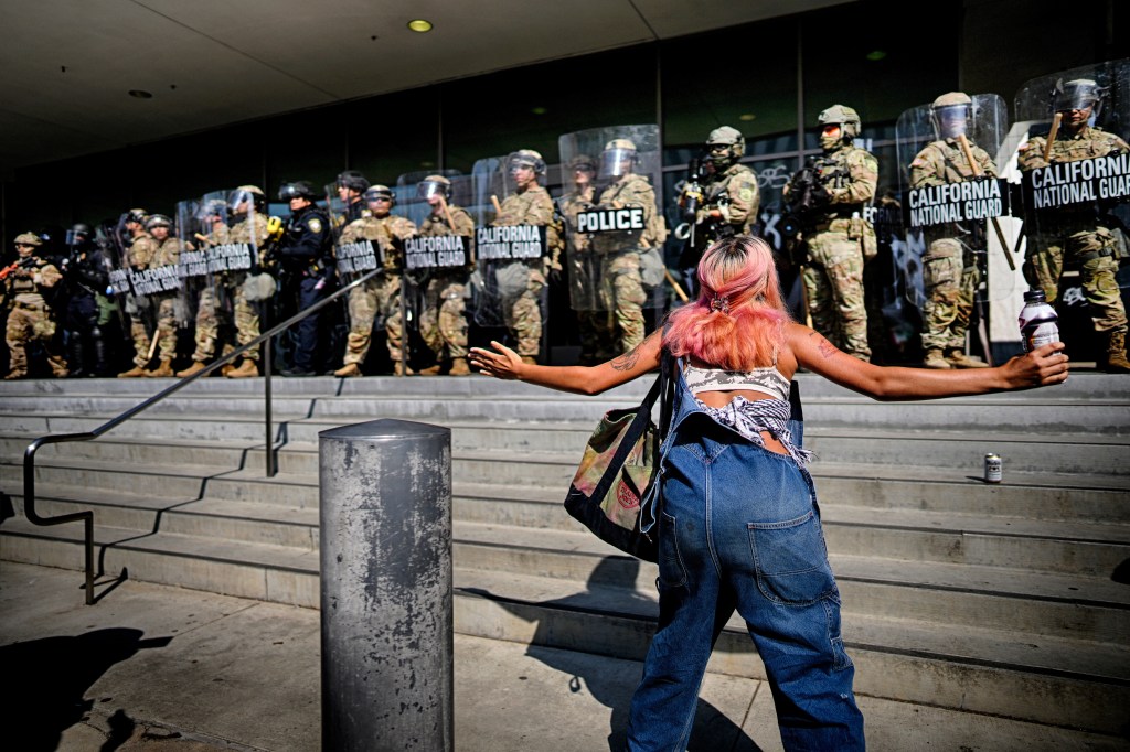 A protester with pink hair, wearing overalls and a bandana, taunts a line of California National Guard and police officers on steps.
