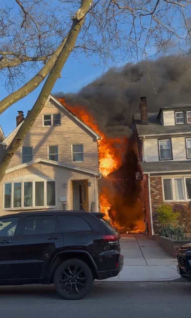 House engulfed in flames with thick black smoke billowing out, next to another house, and a black SUV parked in front.