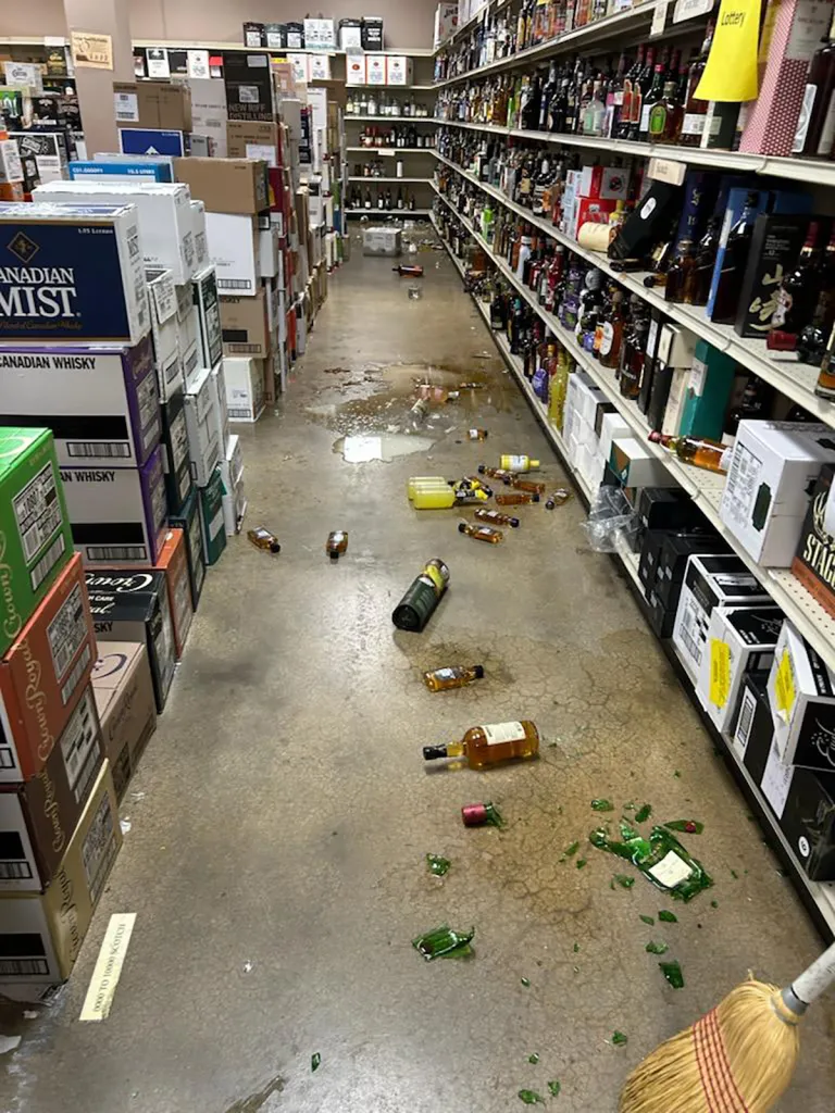 Liquor store aisle with broken bottles, spilled liquor, and debris on the floor.