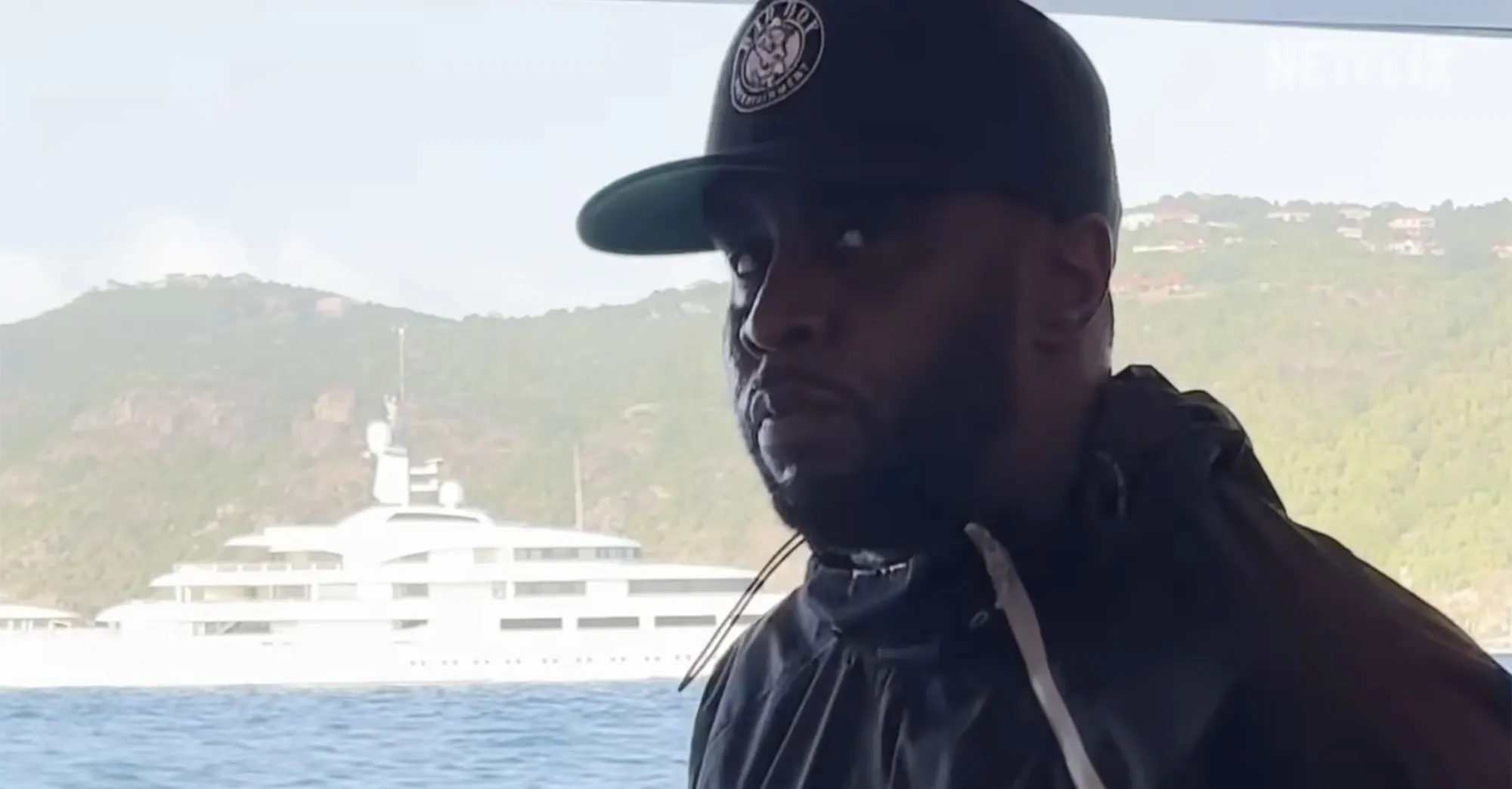 Sean Combs wearing a baseball cap and dark jacket on a boat with a large yacht and hills in the background.