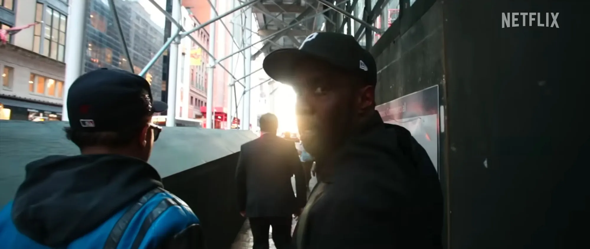 Sean Combs in a black hat looking back, with two other men in front of him, walking under scaffolding in a city.